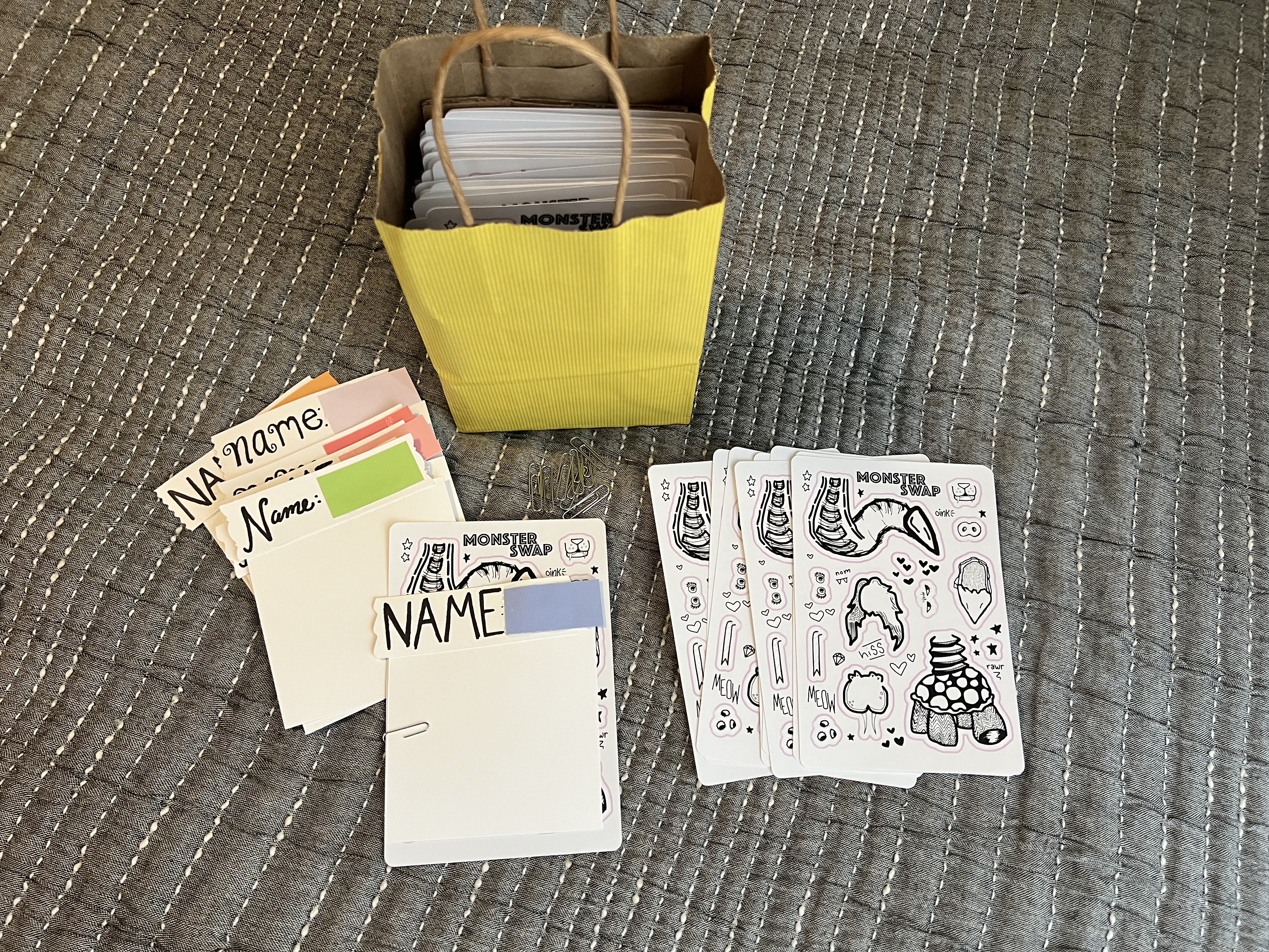 On a bedspread sits a smaller yellow gift bag. In front are two stacks of monster sticker sheet & card pairs