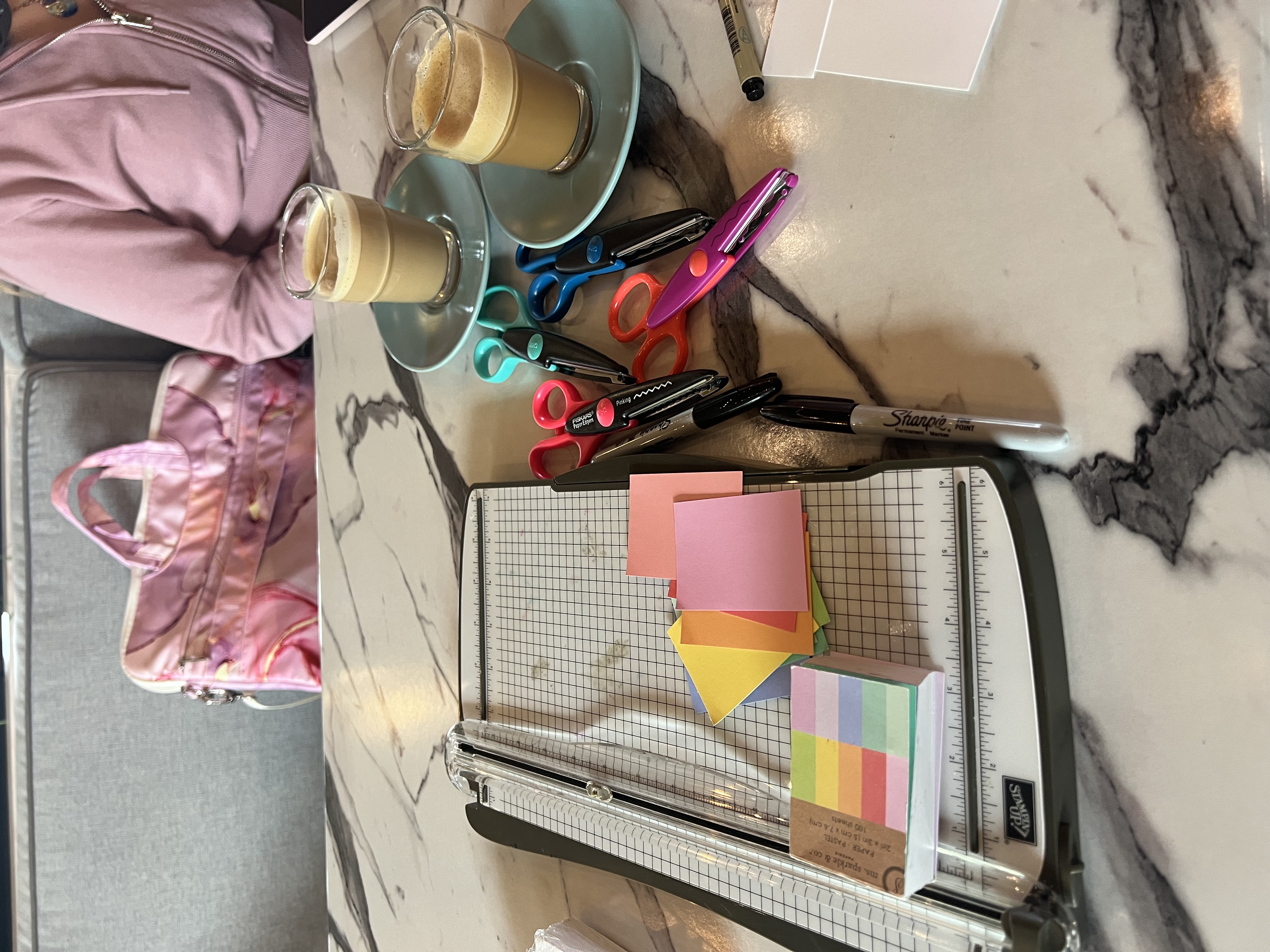 A paper cutter, patterned scissors, and small slips of colorful paper lay on a marbled cafe table. In front sits a small cortado in a for-here cup, and in the back is sitting a blond girl with a pink sweater.