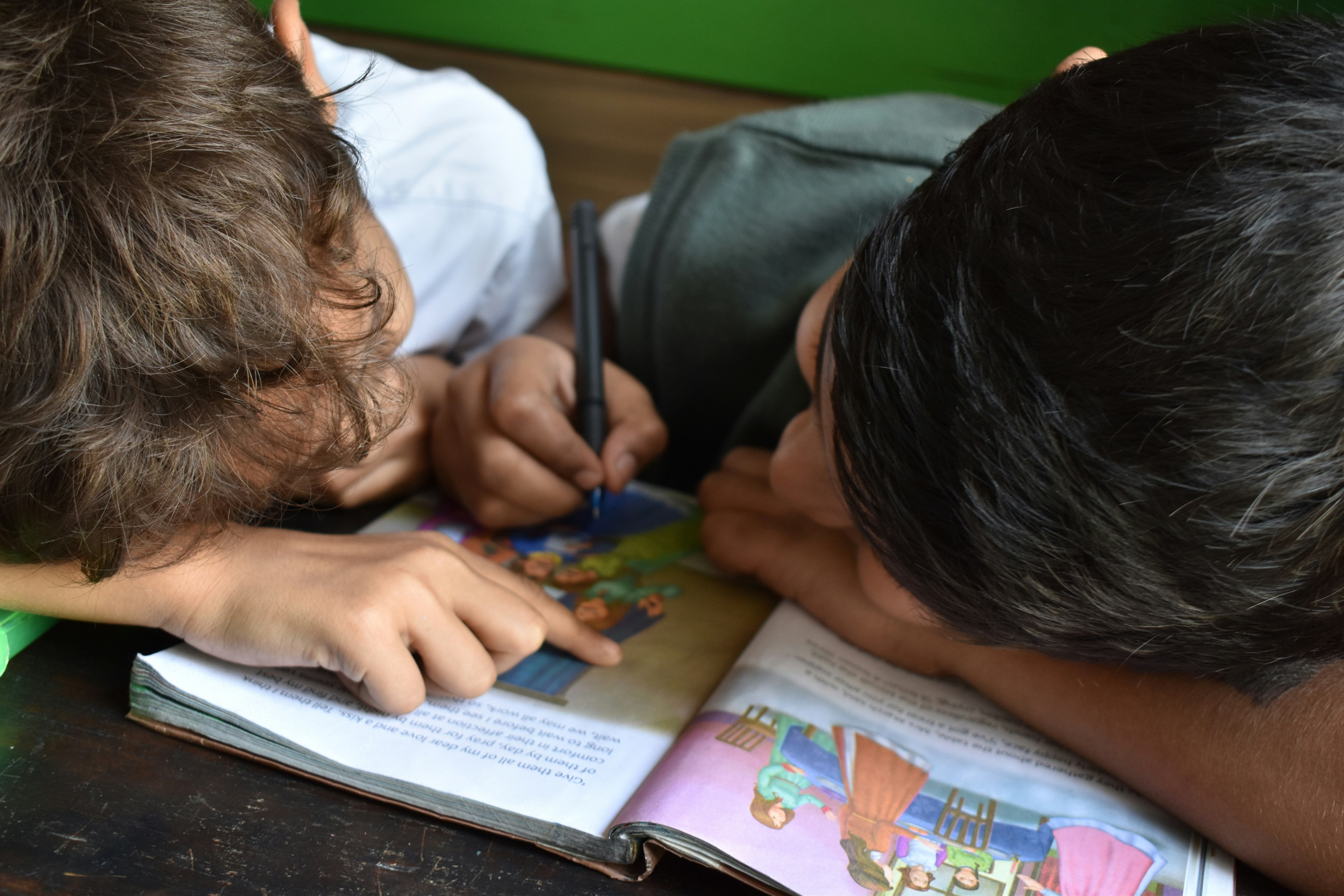 Two children bent over marking,in pen, a schoolbook with pictures and text